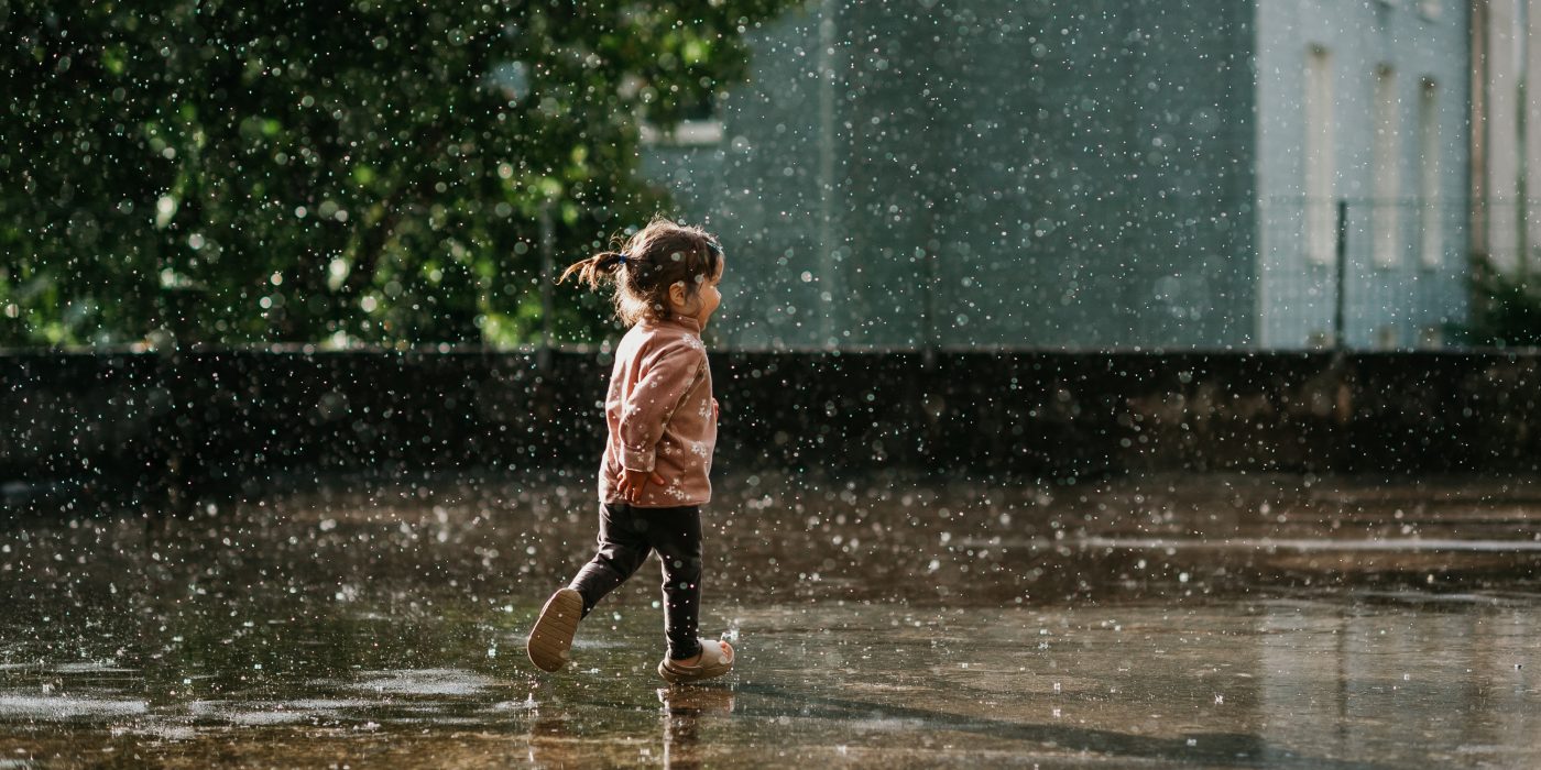A horizontal photo of a cute toddler girl with brown hair and a ponytail in a pink top running and having fun in summer rain in sunshine and raindrops and puddles
