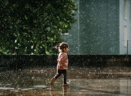A horizontal photo of a cute toddler girl with brown hair and a ponytail in a pink top running and having fun in summer rain in sunshine and raindrops and puddles