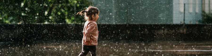 A horizontal photo of a cute toddler girl with brown hair and a ponytail in a pink top running and having fun in summer rain in sunshine and raindrops and puddles